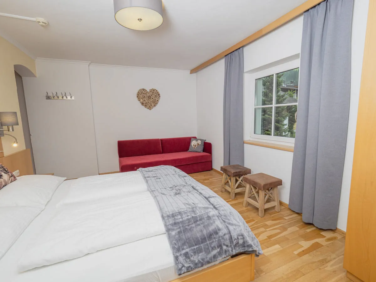 Innenansicht eines Zimmers mit einem Bett, rotem Sofa und Holzstühlen