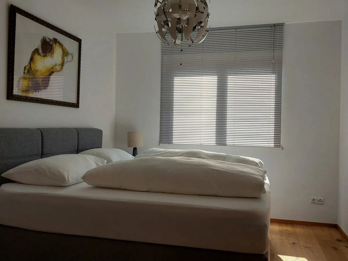 Schlafzimmer mit Bett, Kunstwerk an der Wand und Fenster mit Jalousien.